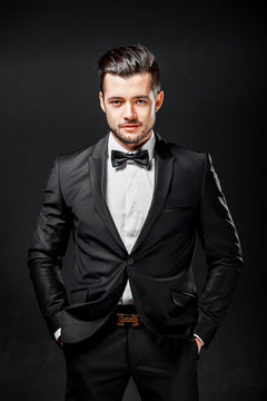 portrait of confident handsome man in black suit with bowtie posing in dark studio background