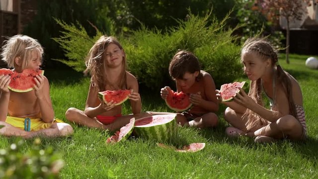 Four kids sitting on grass outdoors eating big watermelon smiling playing in slowmotion