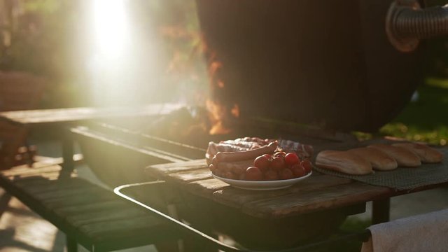 Barbecue grill with burning flame standing near tabel wtih plate of sasuages meat tomatoes and buns outdoors. In slowmotion