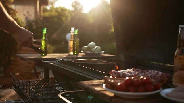Man's hand puts sausages and meat on barbecue grid outdoors while sun shinning in slowmotion