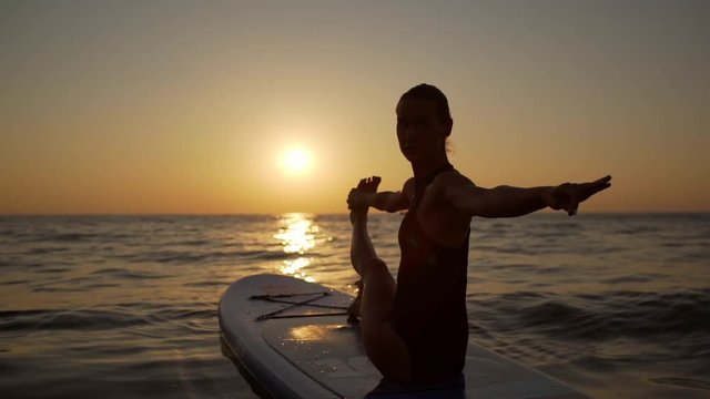Young active woman sitting on supboard and doing yoga asana opposite sun. Silhouette in slowmotion