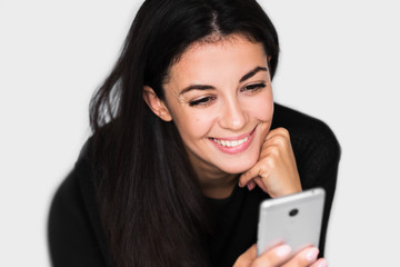 Close up portrait of attractive young happy woman with smart phone with healthy toothy smile,with hand on chin, reading or text message, on light grey background. Technology and business theme.