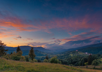 Beautiful summer sunset landscape in Carpathian mountains. Ukraine. Green pasture with wildflowers and shepherds house in the middle ground. Small private hotel as small detail far away.