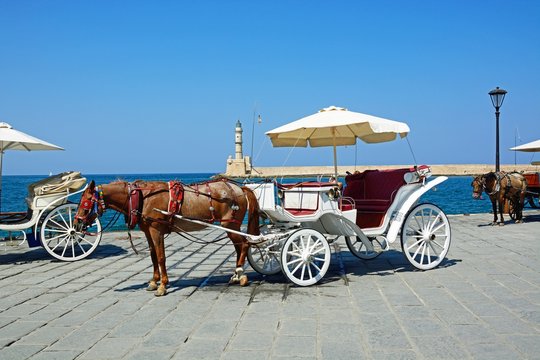 Horse drawn carriages on the quayside with the Venetian lighthouse at the harbour entrance to the rear, Chania, Crete.