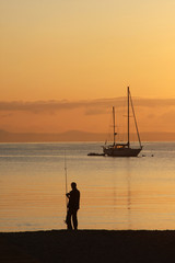 Man fishing in the sea from a beach in early morning sun.