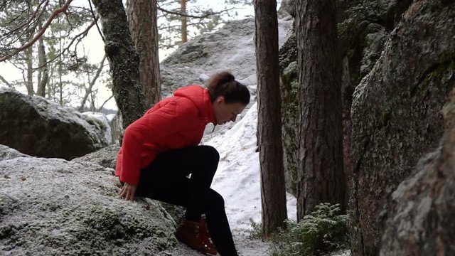 young woman in bright jacket ties the laces on her boots during the hiking through the huge rocks near the frozen lake and pine tree forest