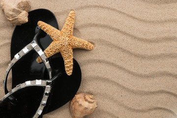 Black flip-flops with rhinestones and seashell, starfish on the wavy sand.