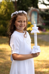 Śliczna dziewczynka w białej sukience trzyma zapaloną świecę.