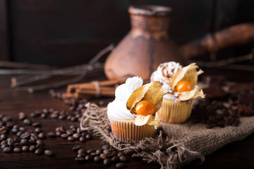 Chocolate cupcakes with white butter cream, decorated with winter cherry on a dark wooden background. Decorated with cinnamon sticks, coffee beans