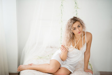 Curly blonde girl in a white silk gown boudoir robe on bed.