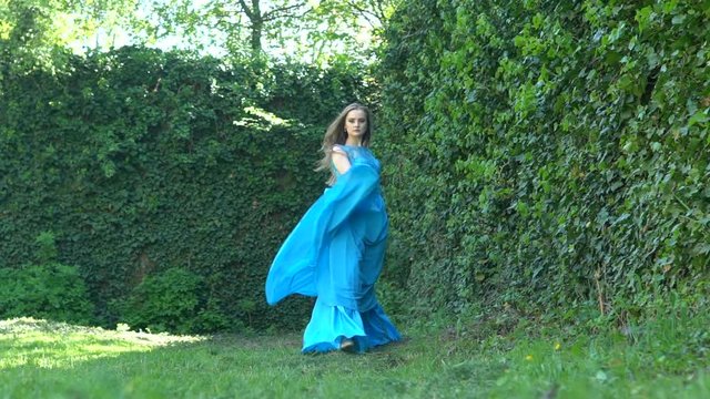 Slow motion: Girl with long hair in a blue dress walk in the green garden. Beautiful gait of a young woman in a long dress