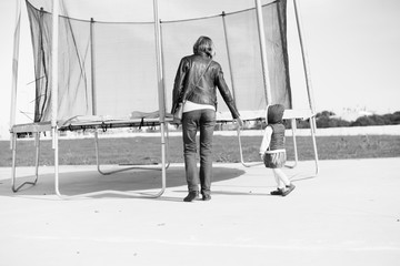 Black and white back view image of parent with child and trampoline. Family time ready for jumping on sunny outdoors background