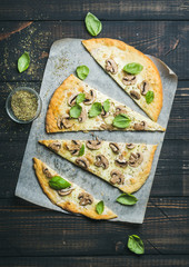 Homemade mushroom pizza with basil cut in slices and spices in glass on baking paper over dark scorched wooden background, top view, vertical composition