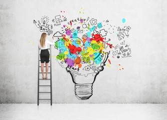 Rear view of a blond woman standing on a ladder and drawing a large and colorful light bulb sketch...