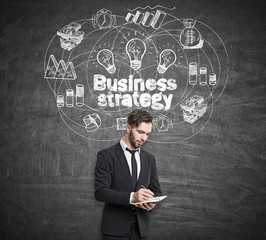 Portrait of a thoughtful businessman with a notebook standing near a blackboard with business strategy drawing on it.