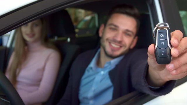 Close up of attractive caucasian man showing car key through the window. Young couple sitting in the car at the dealership. Brunette bearded guy sticking his arm out of the car window
