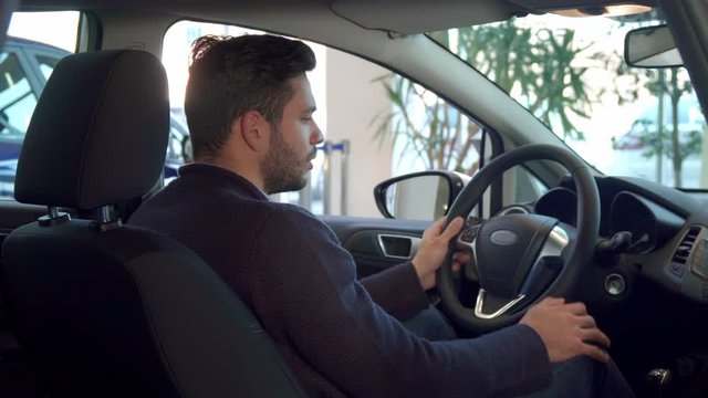 Handsome brunette man showing his thumb up inside the car. Attractive bearded guy approving the car at the dealership. Young male driver turning his face to the back seat