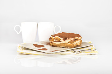 Tiramisu /
Tiramisu cake in a plate with a cocoa decoration and two cups of coffee on a white background