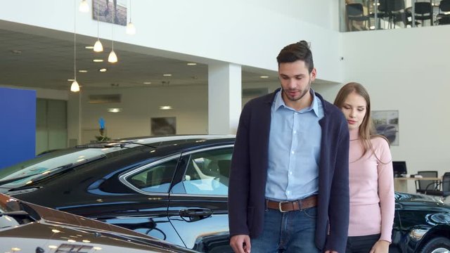 Attractive young couple choosing car at the dealership. Caucasian man and woman standing against background of black sedan. Brunette bearded guy and his pretty girlfrined looking at one of the car