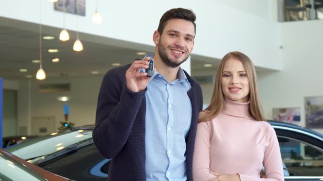 Handsome caucasian man showing the car key at the dealership. Young couple smiling for the camera at the car showroom. Attractive brunette guy standing side by side with his pretty girlfriend against