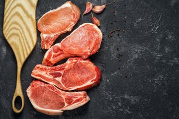 Four large raw pork steaks on the black background with cutting board, garlic and pepper. Copy space for your insctiption.