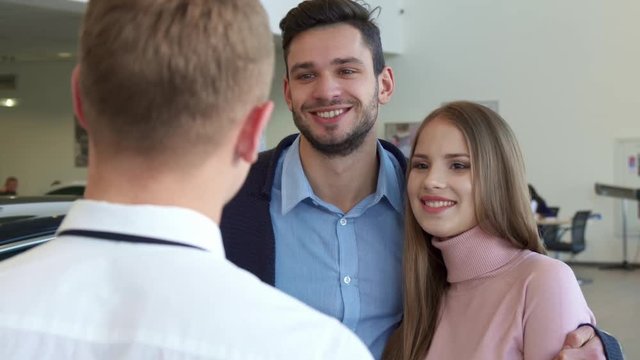 Pretty caucasian woman getting the car key from the salesman at the dealership. Handsome brunette man embracing his attractive girlfriend. Young brunette bearded man and his brown haired girlfriend
