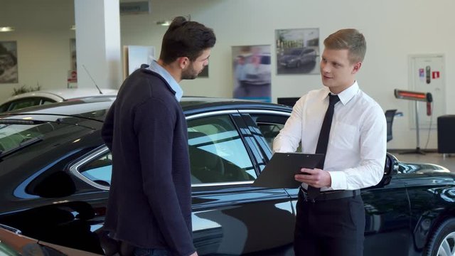 Attractive blond salesman showing something on clipboard to his client. Young male sales manager dealing with brunette bearded man at the dealership. Two caucasian handsome guys shaking hands against