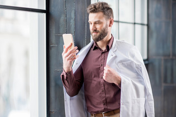 Portrait of handsome confident doctor with medical gown with smart phone standing near the window in the modern dark interior clinic or office