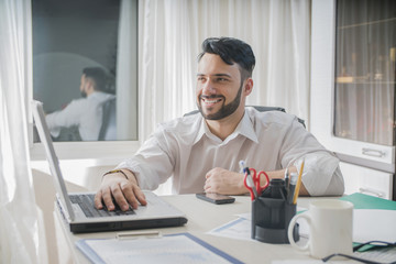 male freelance sitting in an office behind a computer laptop  smiling think and dream