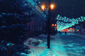 European winter night city street with garlands between the pillars. Lanterns light up on the left before the New Year fir tree