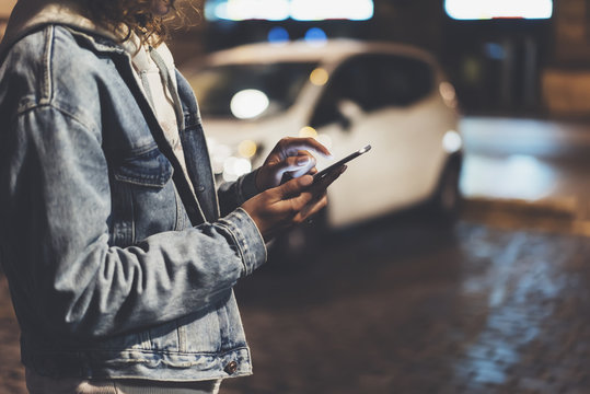 Girl pointing finger on screen smartphone on background auto taxi, illumination bokeh light in night atmospheric city, hipster using and texting mobile phone, mockup glitter street lifestyle