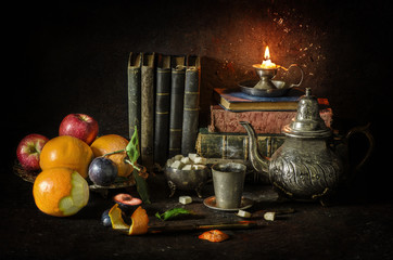 Obraz premium Classic still life with vintage silver plate full of oranges,apples and plums placed with elegant old kettle,sugar,cup of tea,old books and illuminated candle..