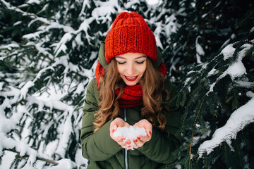 Winter walk. Young girl in red knitted winter hat and scarf holds snow in hands in winter snowy forest