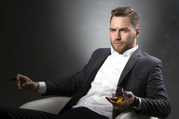 Portrait of chairman sitting in white chair smoking a cigar with cognac over grey background