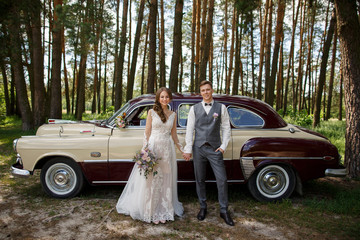 Wedding couple, beautiful bride with rustic wedding bouquet in hand and elegant groom in grey suit with vest holding hands and looking to camera in forest on wedding retro car background