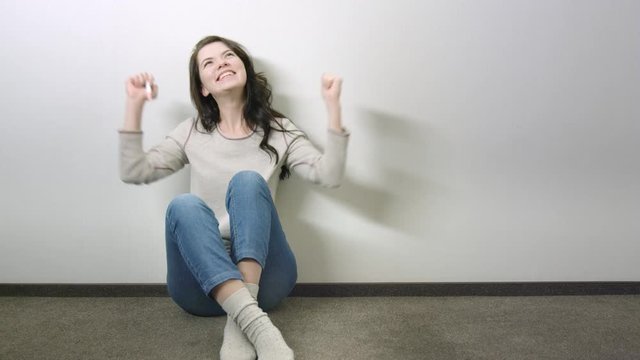 Happy young attractive woman sitting on the floor near the wall and rejoice at positive result of pregnancy test
