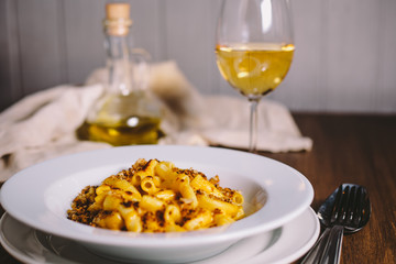 Cheese flavored rigatoni pasta in a white plate on a wooden table with kitchen appliances, glass of dry white wine and glass jug with olive oil on the old gray kitchen towel in the background
