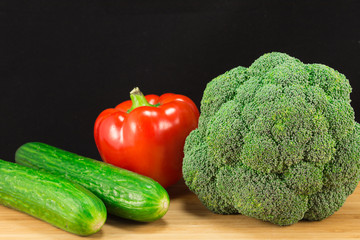 Broccoli, paprika and two cucumbers on the wooden board with black background. Copy space