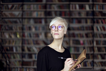 Portrait of woman with blonde hair and eyeglasses in a library, opened book. Hipster student. Education concept