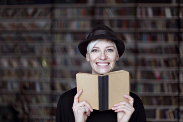 Portrait of woman in black hat with opened book smiling in a library, blonde hair. Hipster student girl