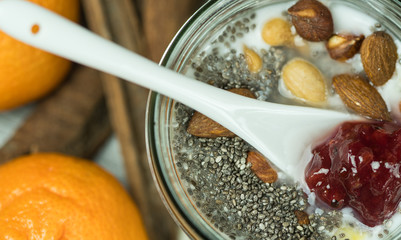 Macro shot of a white ceramic teaspoon of strawberry jam dipped in healthy breakfast mix, almonds, hazelnuts and walnuts chia seeds and cereal. Tangerines and Cinnamon sticks in the background.