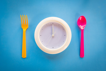 white clock on coloured background, at dinner spoon and fork waiting for food