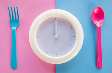 white clock on coloured background, at dinner spoon and fork waiting for food