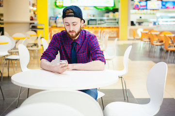 portrait young man with a beard in a plaid shirt and a tie and a cap, sits on a chair at a table food court mall