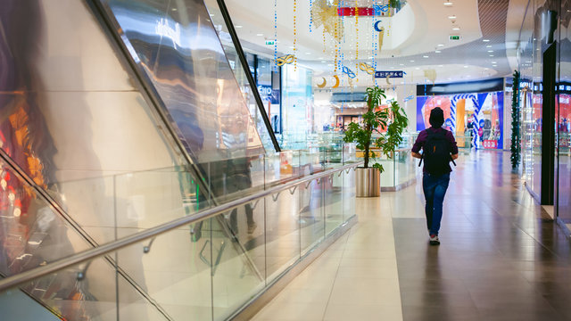 young man with a beard in a plaid shirt and a tie and a cap, walking through the mall on a background of shops and escalator