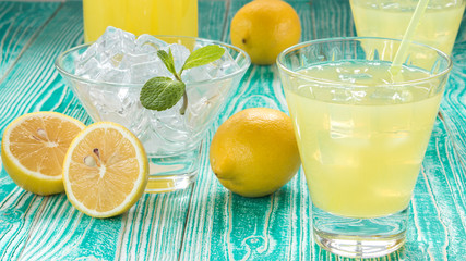 lemonade or limoncello in a yoke stopper bottle, drink in glasses, sherbet glass with ice cubes decorated by mint leaf, lemon fruits on turquoise colored wooden table 