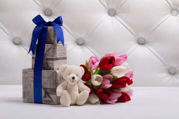 Teddy bear sits near the bouquet of tulip and gift boxes with newspaper texture. Toy and flowers closeup. Background for a postcard.
