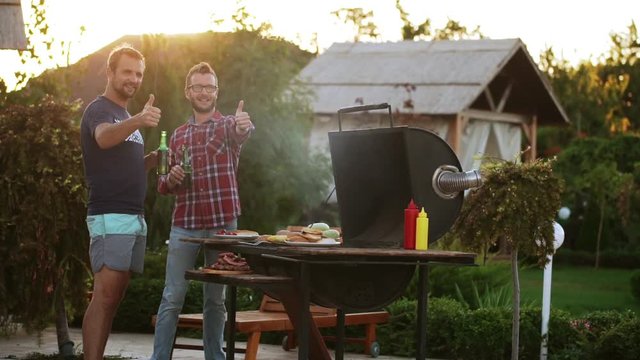 Two male friends drinking beer while making barbecue and showing ok with thumbs in slowmotion