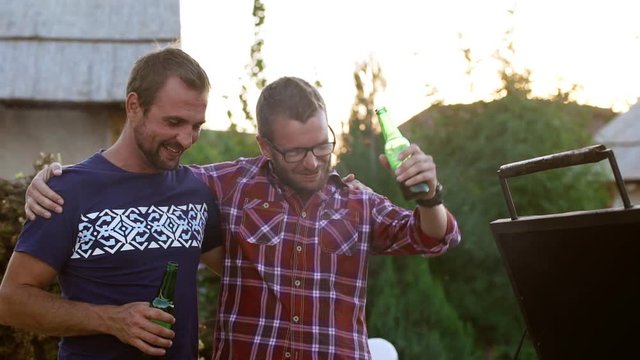 Two male friends huging each other shoulders standing near barbecue grill drinking beer talking smiling laughing in slowmotion