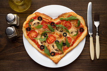 Tasty hot pizza in heart shape with chicken and mushrooms and cutlery on wooden table.